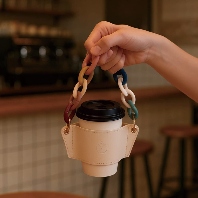 A drink bottle holder made of cream leather with a multicoloured acrylic strap, holding a coffee cup.
