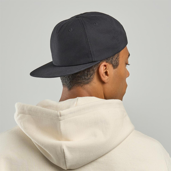 A black cap with a flat peak is worn backwards by a person in a cream-coloured hoodie. The cap has a logo.