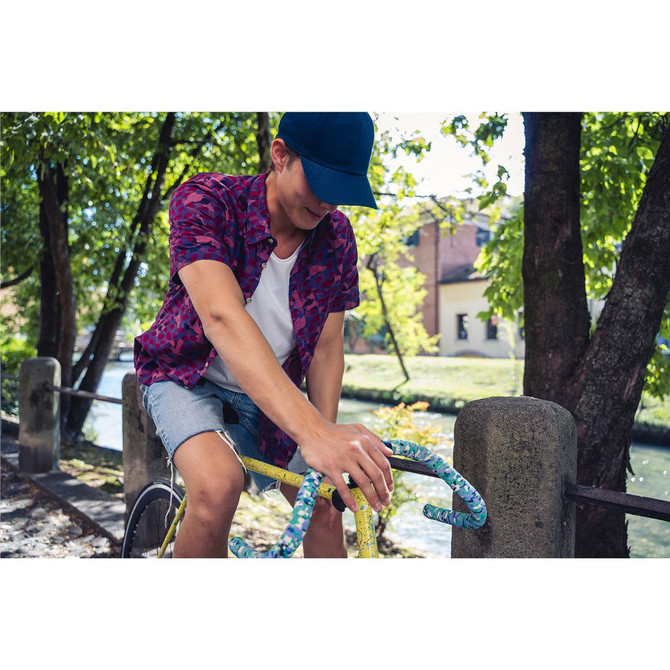 A person wearing a navy cap rides a bicycle along a riverside path, surrounded by greenery.