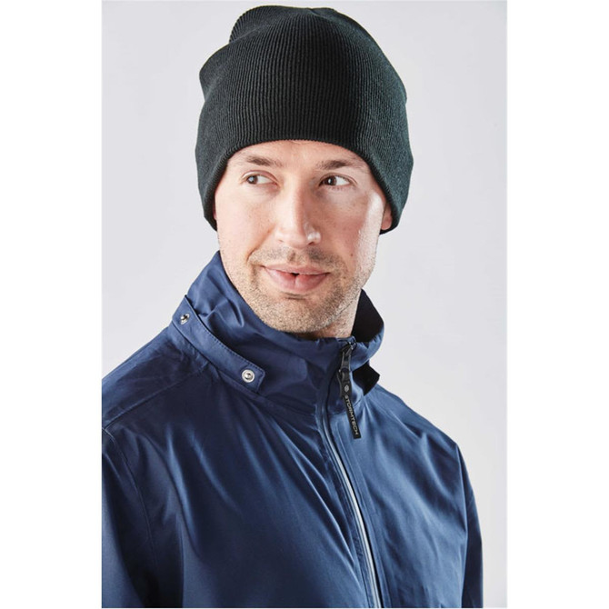 A dark green knit beanie worn by a man in a blue jacket, featuring a logo on the beanie.