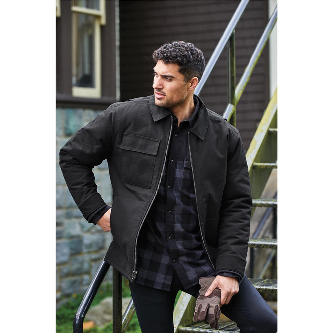 A men's flatiron work jacket in black, featuring a front pocket and worn with gloves. The model is standing on stairs.