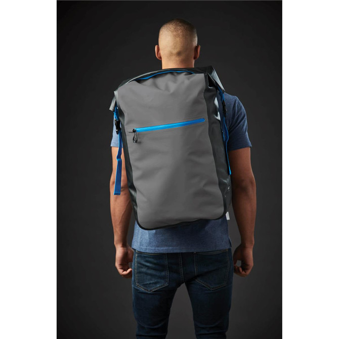 A grey backpack with blue accents is worn on the back of a person. The backpack features a front zip pocket and has a logo.