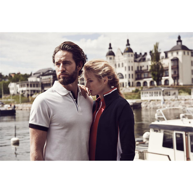 A man in a white polo shirt with black accents stands next to a woman in a dark jacket, near a waterfront.