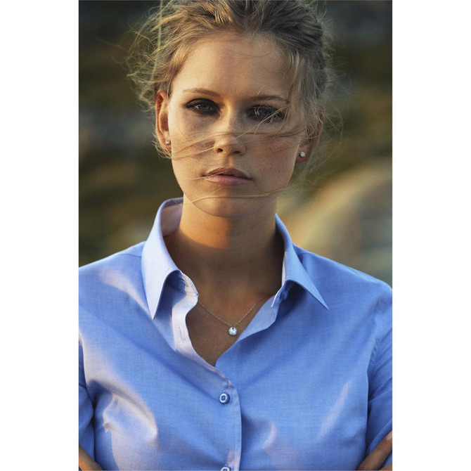 A woman wearing a light blue button-up blouse, with soft hair flowing in the breeze, and subtle jewellery.