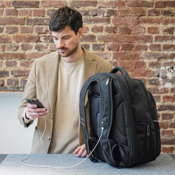 A black backpack sits on a table next to a man using his phone, with a charging cable connected to the bag.