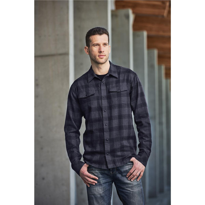 A man wearing a black and grey checked button-up shirt, standing against a concrete wall. The shirt has a logo.