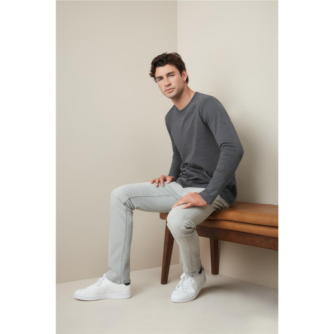 A man in a grey long sleeve T-shirt sits on a wooden bench, wearing light grey pants and white sneakers.