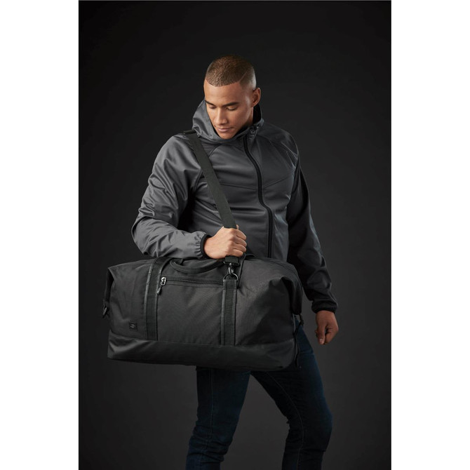 A duffle bag in black is held by a person wearing a grey jacket against a dark background. The bag features a logo.