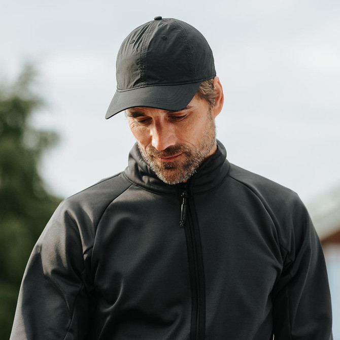 A black cap worn by a man in a black jacket, with a blurred outdoor background. The cap features a logo.