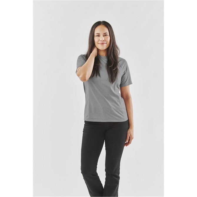 A woman in a grey t-shirt poses against a plain background, wearing black pants. The shirt has a logo.