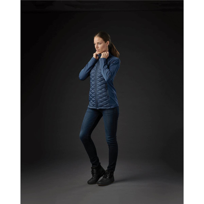 A woman stands wearing a navy thermal shell jacket with a quilted design, paired with dark jeans and boots.