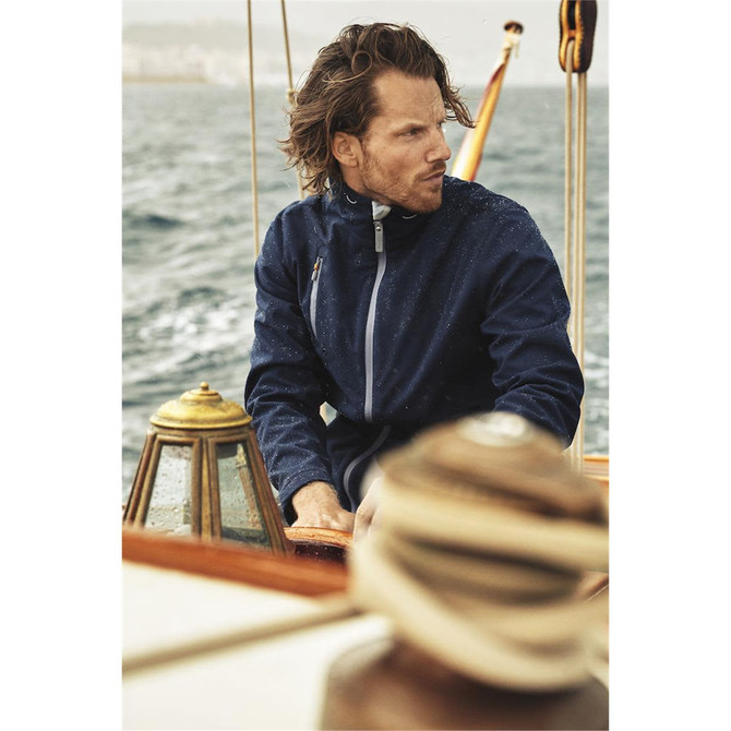 A navy softshell jacket is worn by a man sailing on a boat, with water and a coastline in the background.