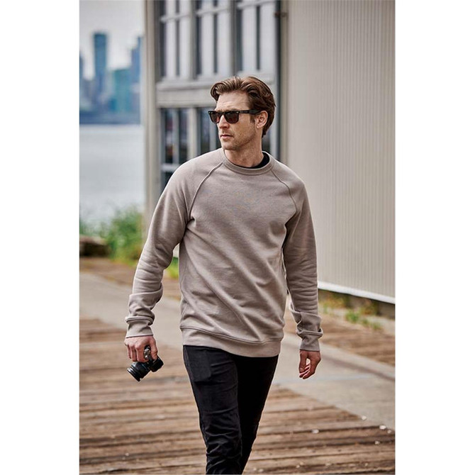 A man wearing a grey crew neck fleece sweater walks outdoors, holding a camera, with a city skyline in the background.