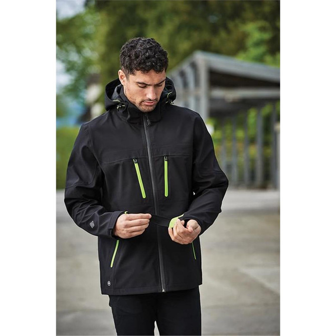 A black softshell jacket with green accents, featuring a hood and zippered pockets. It has a logo on the sleeve.