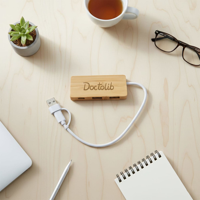 A bamboo USB hub with a white cable, placed on a wooden surface alongside a cup, notebook, and glasses.
