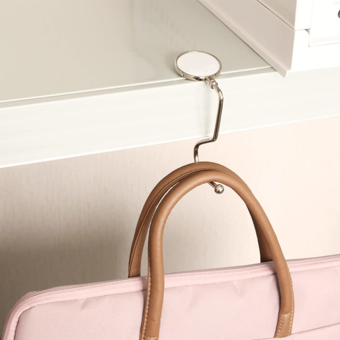 A round, curved metal desk hook in silver holds a pink bag on the edge of a white desk.