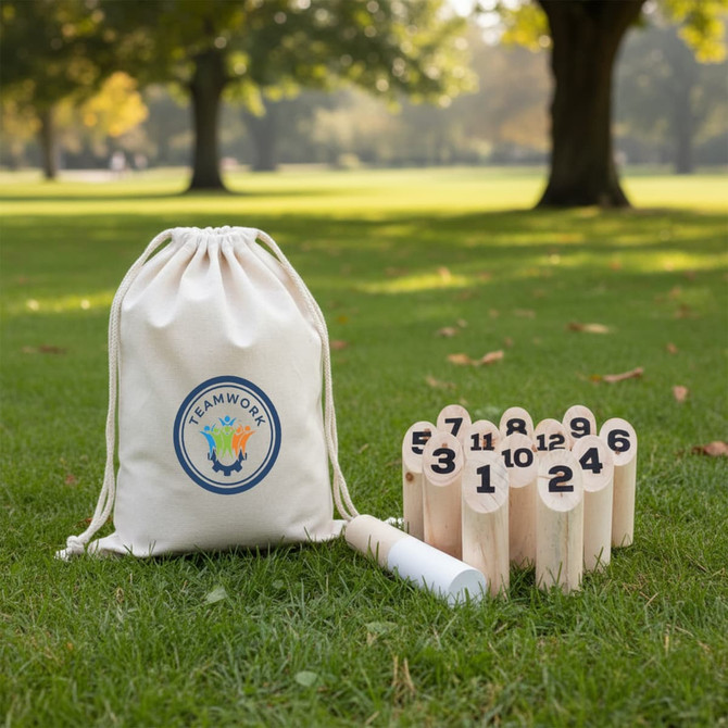A drawstring bag with a logo beside wooden skittle pins numbered 1 to 12, set on green grass in a park.