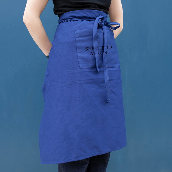 A blue long waist apron with a front pocket and tied at the waist, displayed against a blue background.