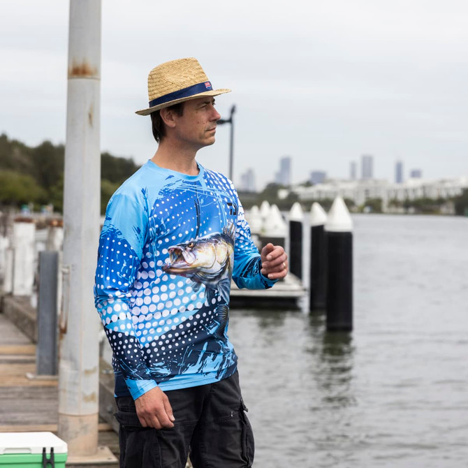 A man wearing a blue long sleeve polyester fishing tee stands by the water, surrounded by a city skyline and fishing gear.