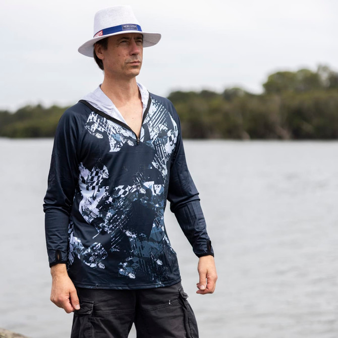 A man in a navy and white patterned polyester fishing hoodie stands by the water, wearing a white hat.
