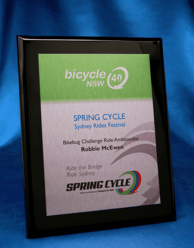 A black plaque featuring a brushed silver printed metal plate, displaying event details and a logo.