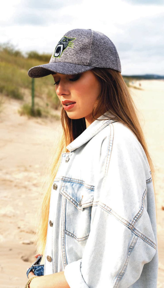 A woman wears a grey cap and a denim jacket, standing on a sandy beach with grass in the background.