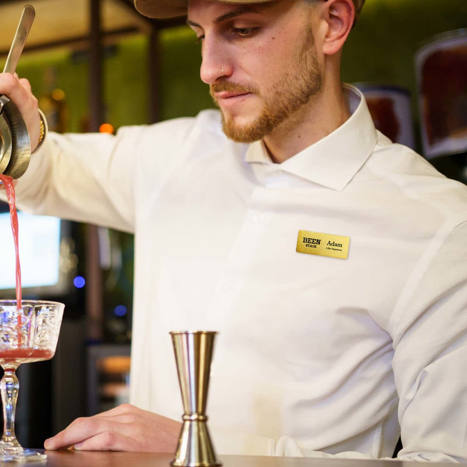 A man in a white shirt pours a drink into a glass, wearing a metal name badge and a hat.