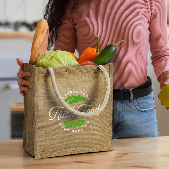 A jute tote bag in natural colour with sturdy handles, filled with fresh vegetables and a baguette.