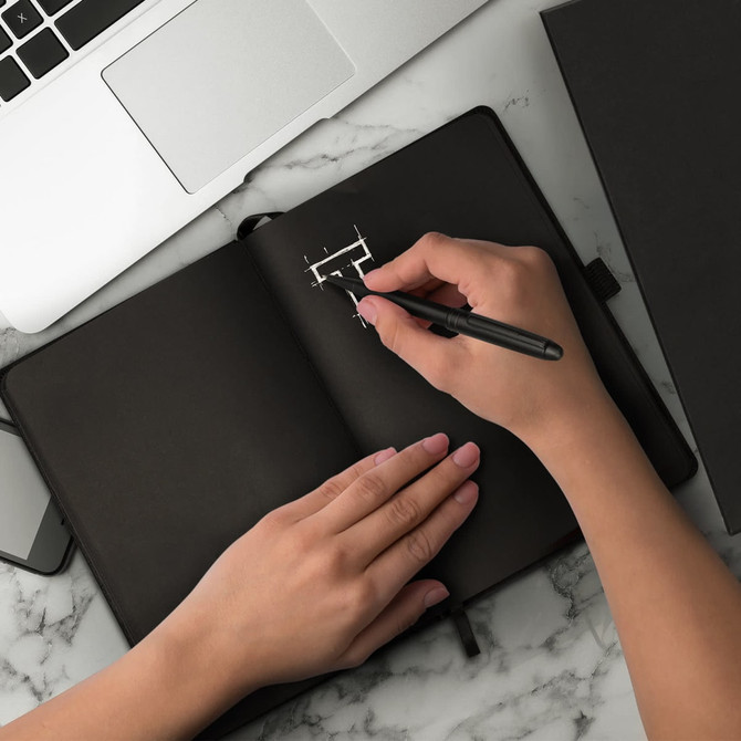 A black notebook with a hand writing inside it, accompanied by a black pen on a marble desk with a laptop.