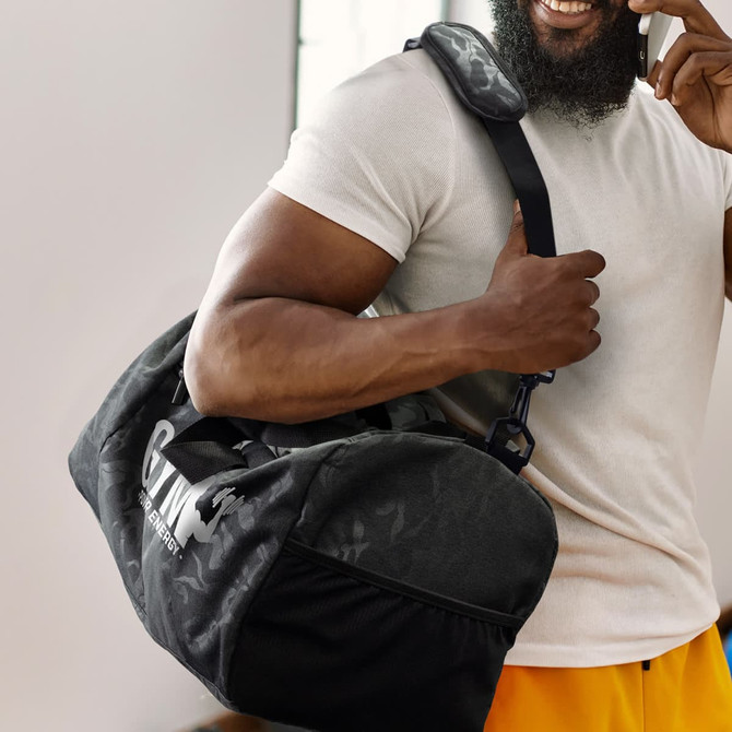 A duffle bag in urban camo pattern, featuring black and grey tones, with a shoulder strap held by a person.