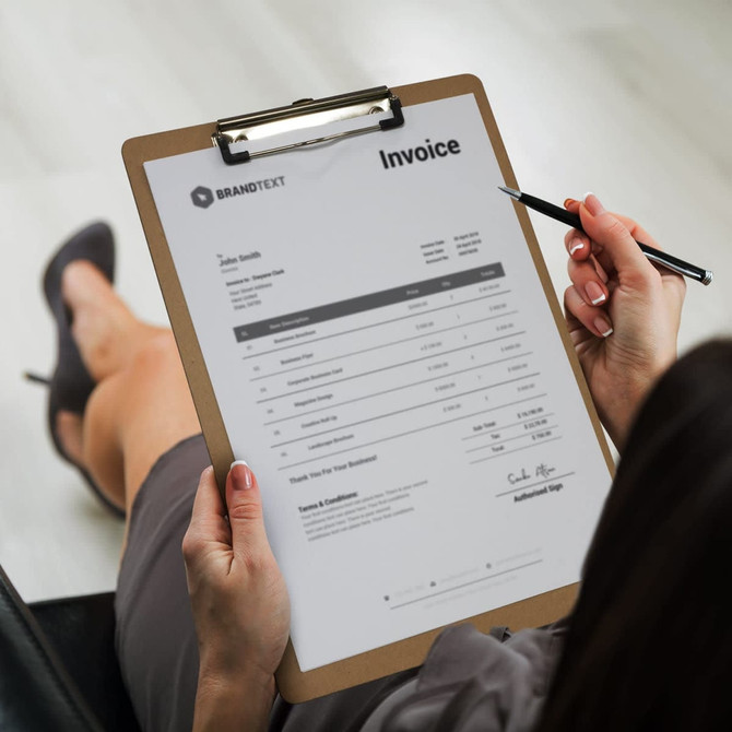 A wooden clipboard holds an invoice with a logo at the top, held by a person writing with a pen.