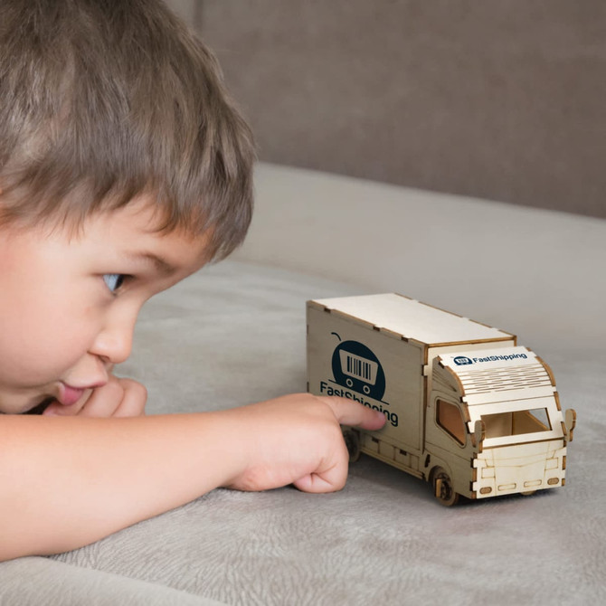 A wooden model of a small truck in natural wood tones, with a logo, alongside a child pointing at it.