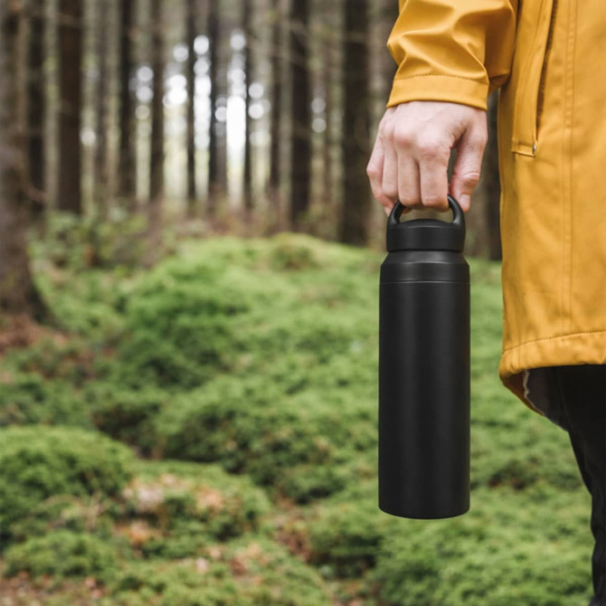 A drink bottle in matte black, featuring a handle, held by a person in a yellow jacket amidst a forested background.