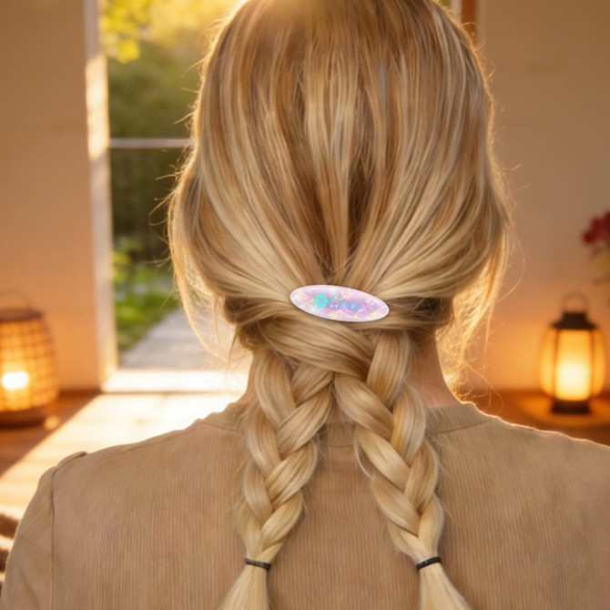 An oval iridescent alligator hair clip holds two braided sections of blonde hair. The setting features warm, ambient lighting.