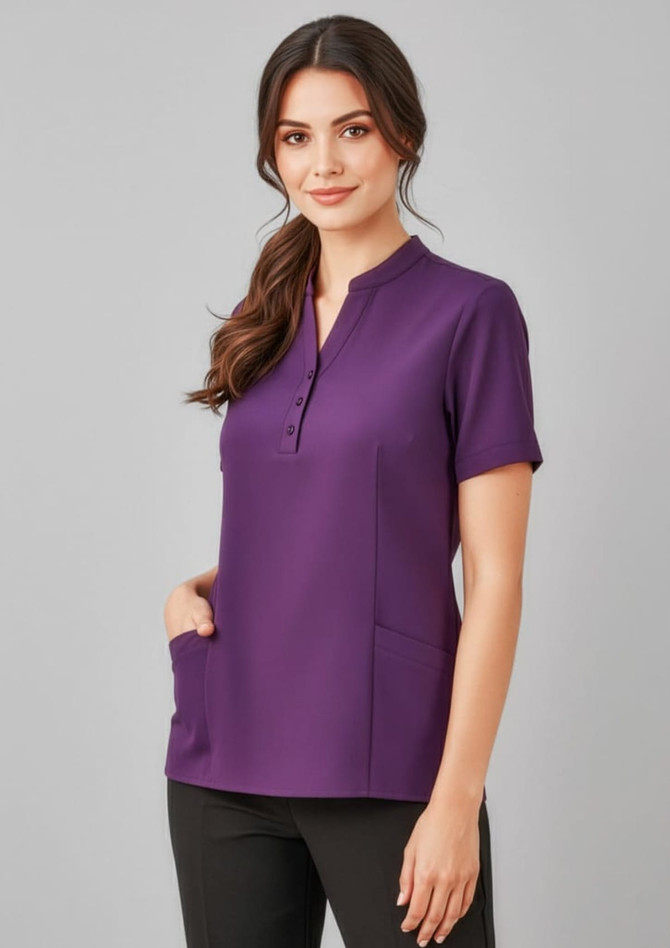 A women's tunic in a rich purple colour with short sleeves and pockets, featuring a button-up placket and a logo.