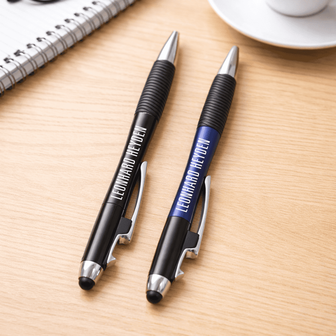 A pair of pens, one black and one blue, with grips, resting on a wooden surface near a cup and notebook.