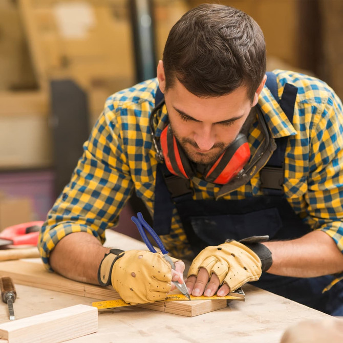 A man wearing a checkered shirt and gloves is measuring and marking wood in a workshop setting.