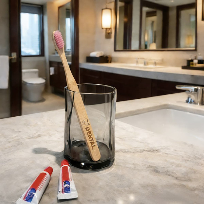 A large flat handle toothbrush with a pink bristle head in a glass, beside two tubes of toothpaste on a bathroom countertop.