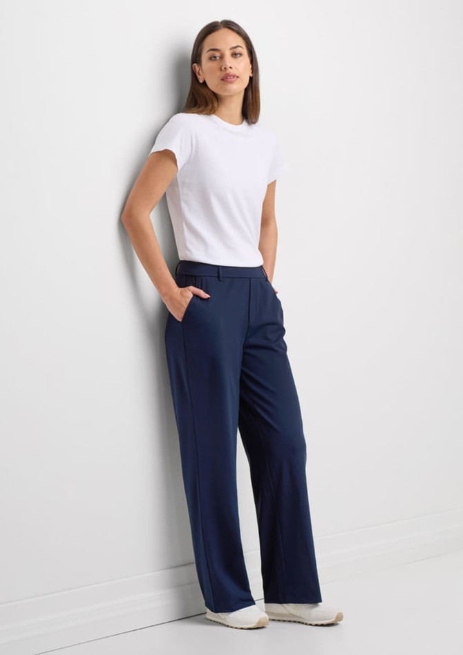 A woman stands against a wall wearing navy blue soft stretch pants and a fitted white t-shirt, with pockets visible.