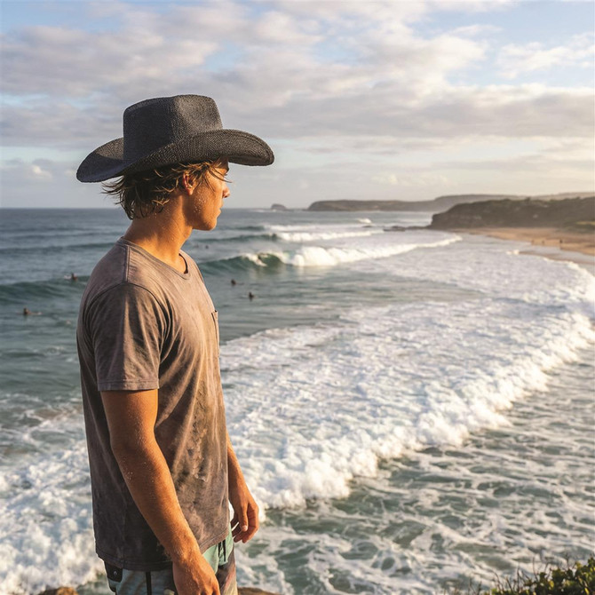 A person wearing a black straw hat stands by the ocean, gazing at the waves and surfers.