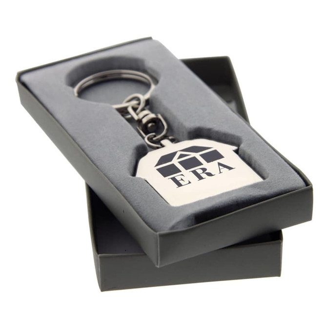 A silver keychain in the shape of a house, presented in a grey box with a logo.