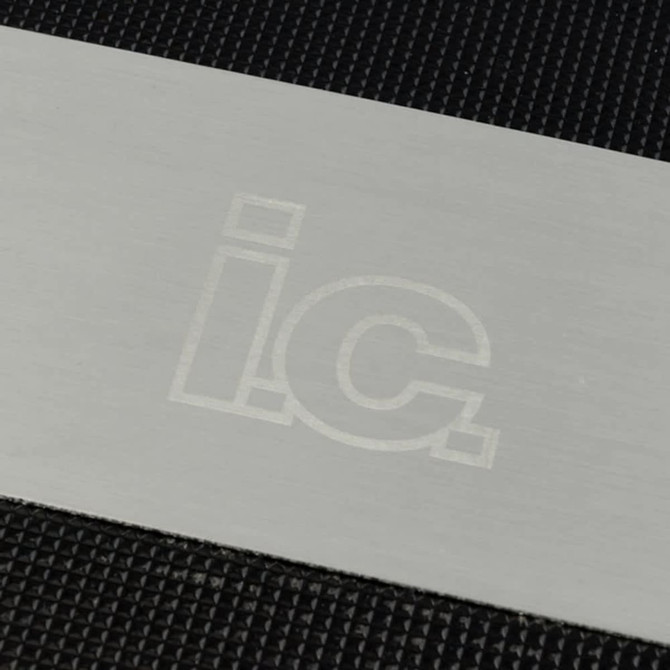 A stainless steel grill spatula is displayed on a textured black surface, featuring a subtle logo.