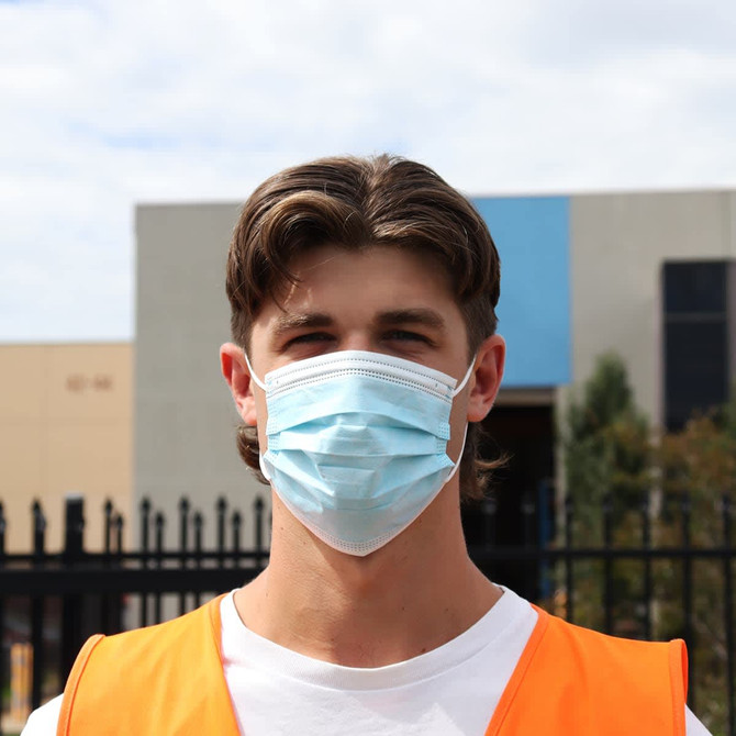 A blue triple-layered cloth face mask worn by a person in an orange vest. The background features buildings and a fence.