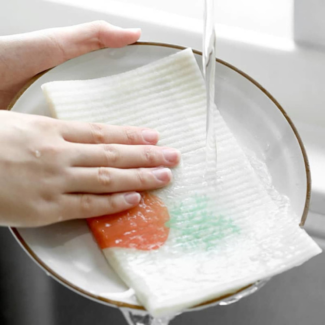 A hand holds a biodegradable dishwashing cloth on a plate under running water, featuring green and orange designs.