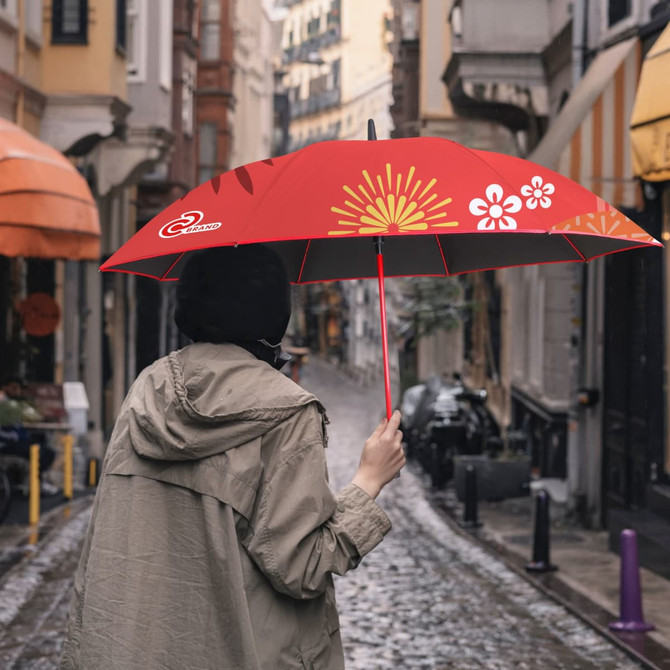 A red umbrella with floral patterns is being held by a person wearing a light coat, standing in a cobblestone street.