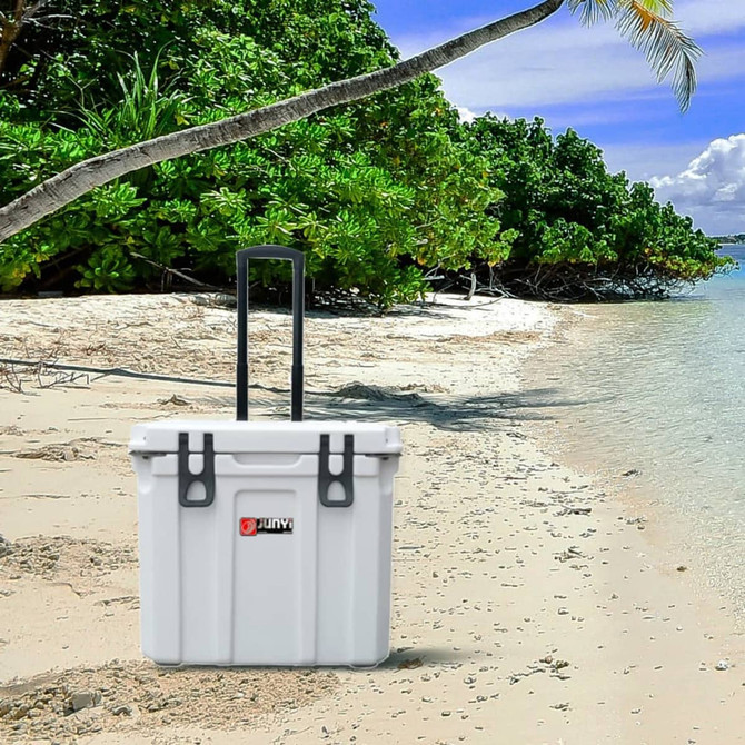 A white cooler box with wheels stands on a sandy beach surrounded by greenery and water. It has a logo.