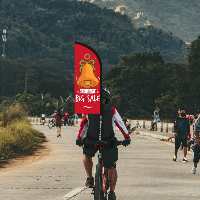 A Feather Backpack Flag features a vibrant red design with a festive bell illustration, set against a hilly background.