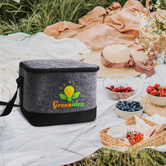 A grey felt cooler bag sits on a picnic blanket, surrounded by food, flowers, and picnic items. It features a logo.