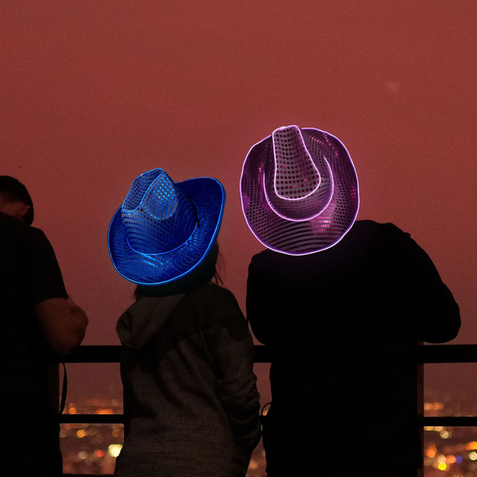 Light-up sequin cowboy hats in blue and pink, worn by two people against a city skyline at sunset.