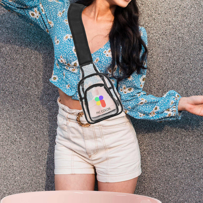 A transparent PVC crossbody backpack with a black strap, held by a person in a blue floral top and white shorts.